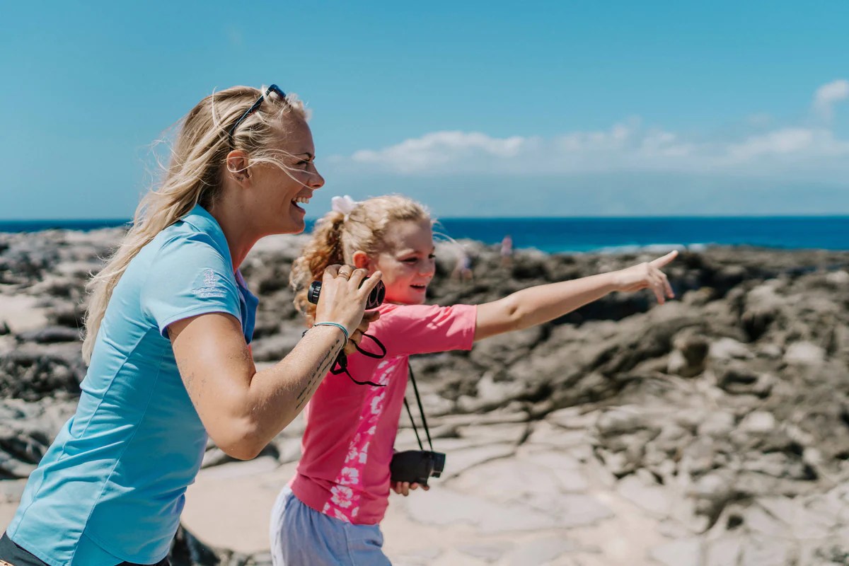 Woman and girl smiling, pointing on rocky beach under clear sky.